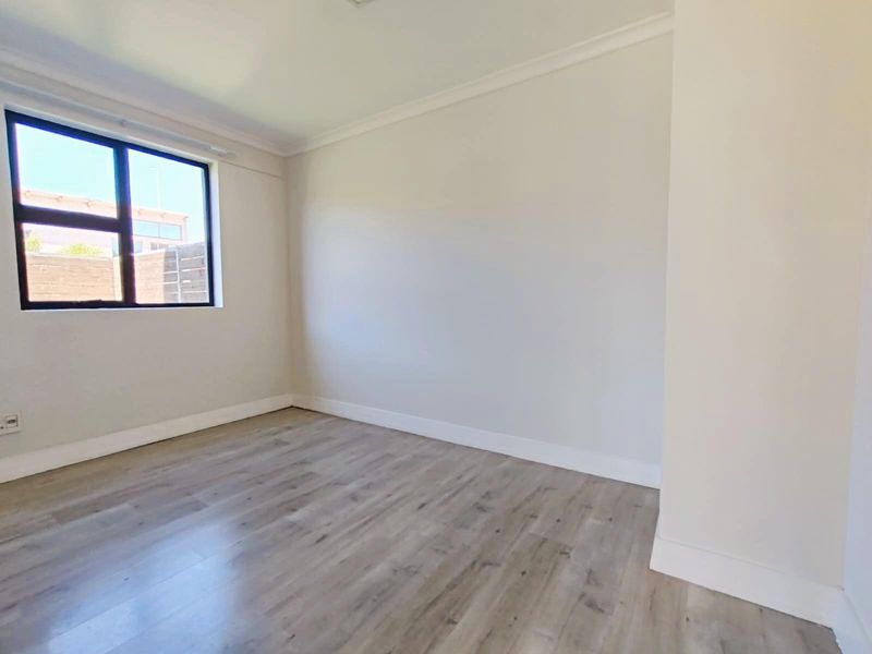 A bright and neutral bedroom featuring modern wood-look laminate flooring, crisp white walls, and a large window providing natural light.