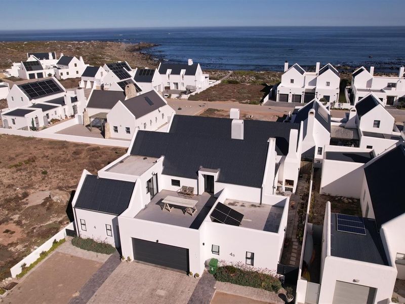 Aerial view of a modern coastal residential estate featuring white-walled homes with dark metal roofs, solar panels, and paved driveways near the ocean.