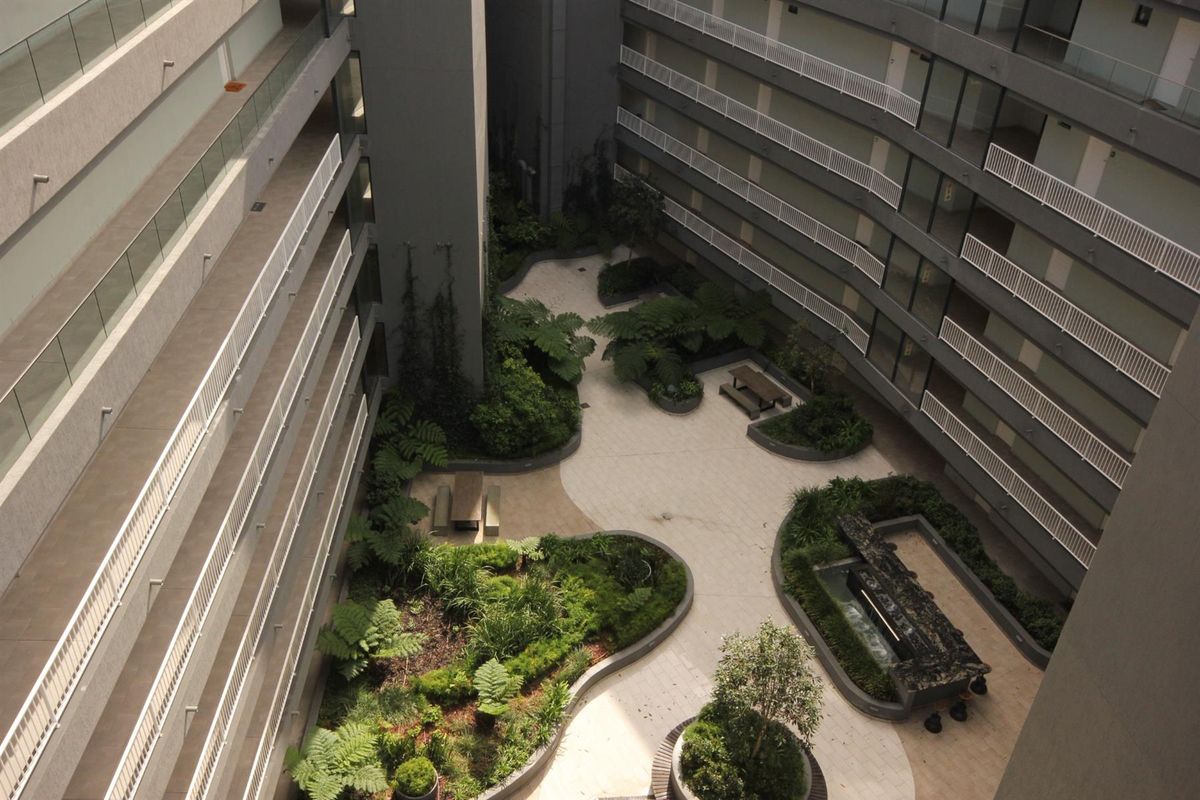 An elevated aerial view of a modern apartment complex courtyard featuring landscaped gardens, paved pathways, and multi-level balconies with glass balustrades.