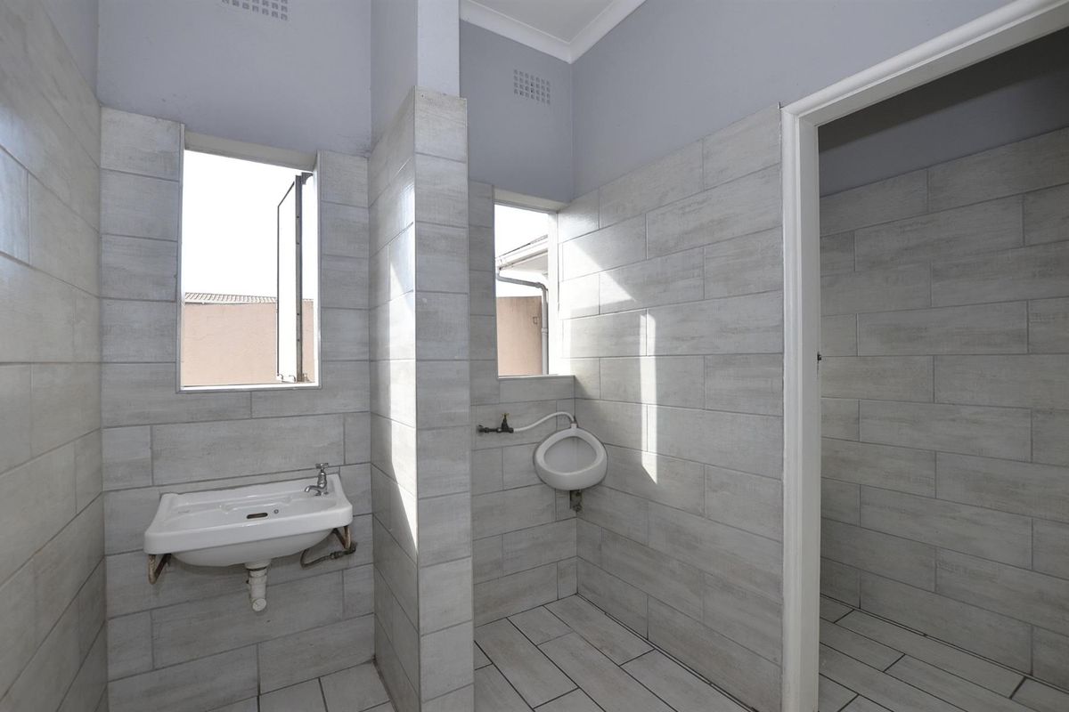 A functional commercial bathroom featuring tiled walls and floors, a wall-mounted basin, and a wall-mounted urinal with natural light from casement windows.