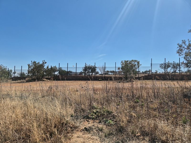 A view of a vacant residential plot featuring a stone retaining wall and perimeter fencing against a clear blue sky.