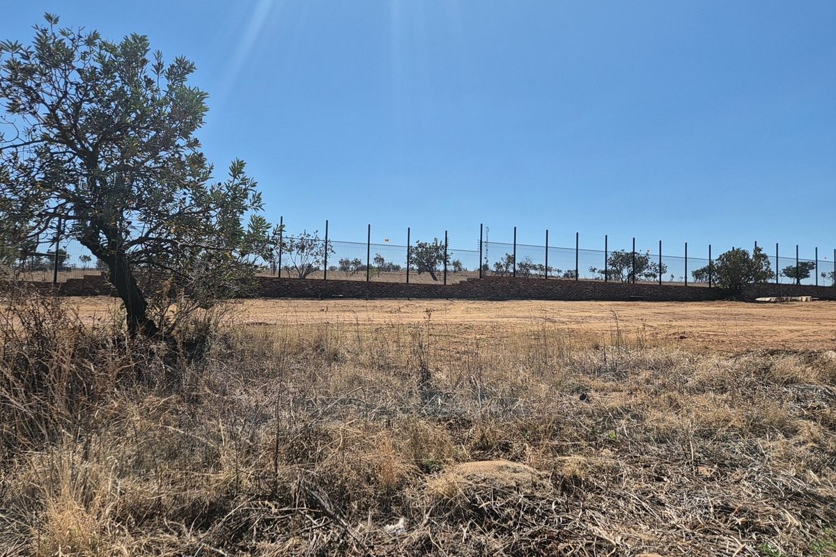 A view of a vacant residential plot featuring a perimeter fence and a low brick retaining wall against a clear blue sky.