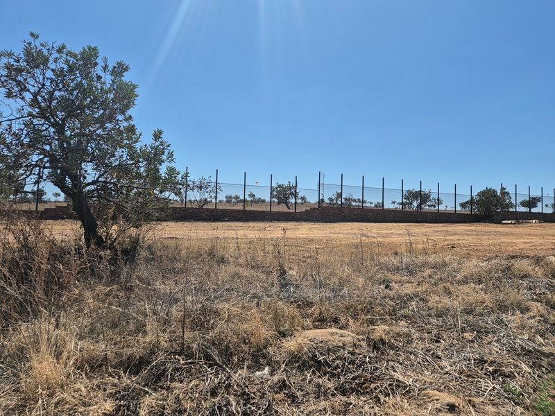 A view of a vacant residential plot featuring a perimeter fence and a low brick retaining wall against a clear blue sky.