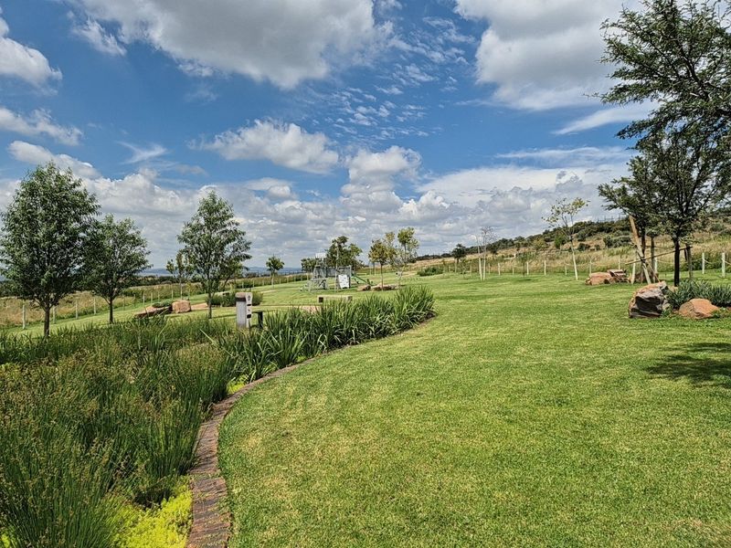 A well-maintained residential garden featuring manicured lawns, established trees, and neat brick-edged garden beds under a clear blue sky.