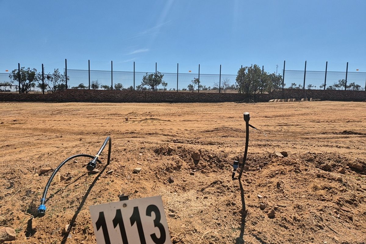 A vacant residential plot featuring a perimeter fence, brick retaining wall, and exposed irrigation infrastructure on a clear, sunny day.