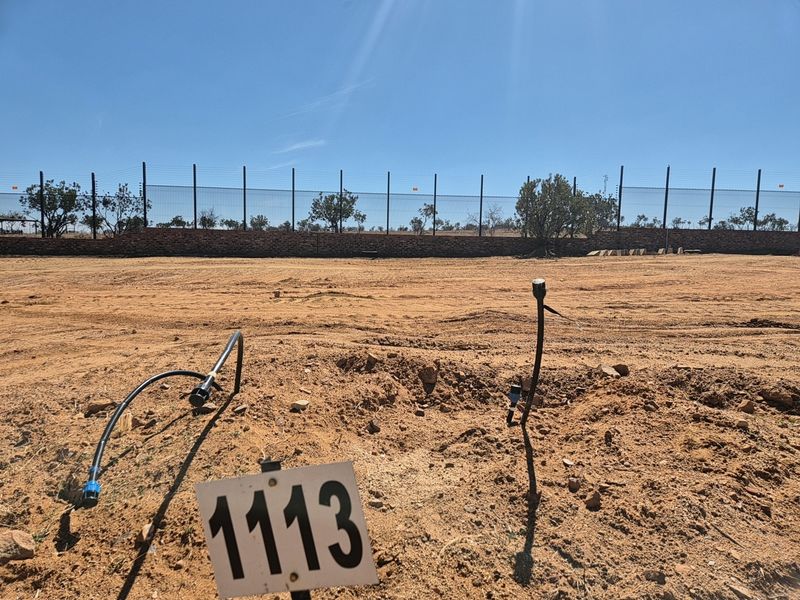 A vacant residential plot featuring a perimeter fence, brick retaining wall, and exposed irrigation infrastructure on a clear, sunny day.