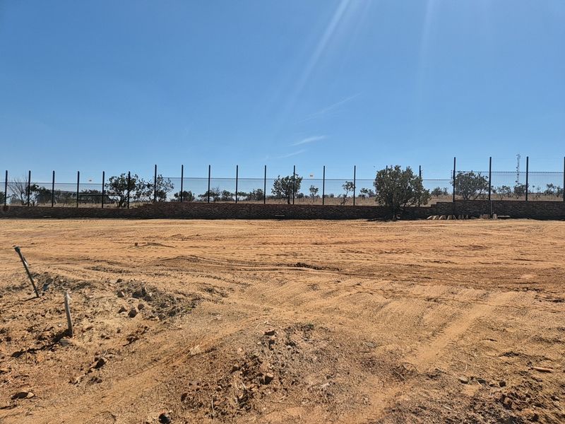 A vacant residential plot featuring a level sandy surface, enclosed by a perimeter fence and a low retaining wall.