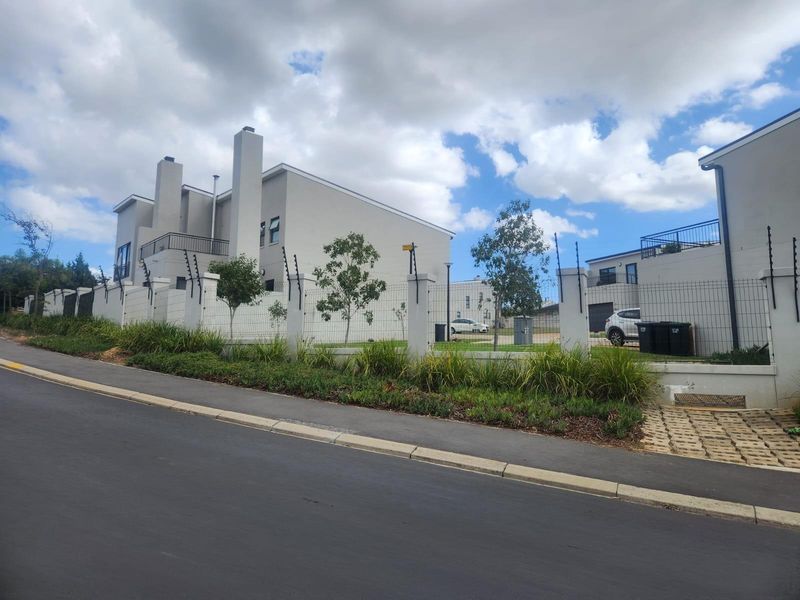 Street view of a modern residential property featuring a secure perimeter wall with electric fencing and paved driveway access.