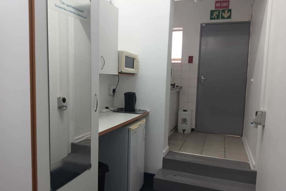 A functional office kitchenette area featuring built-in cabinetry, laminate countertops, and a stepped transition to a rear utility space with an emergency exit.