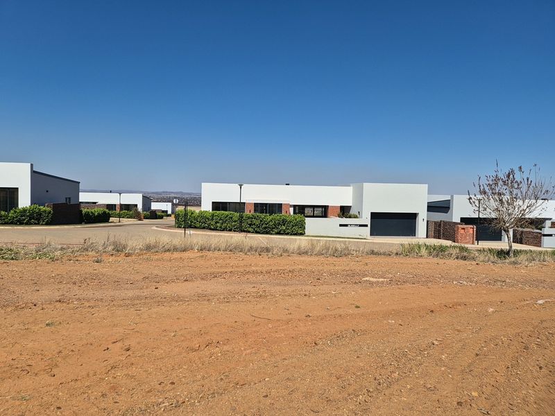 A view of a vacant residential plot looking towards modern, well-maintained homes in a suburban estate setting.