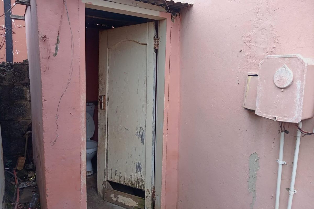 Exterior view of a small, detached outdoor toilet facility with visible wear and tear on the door and surrounding wall structure.