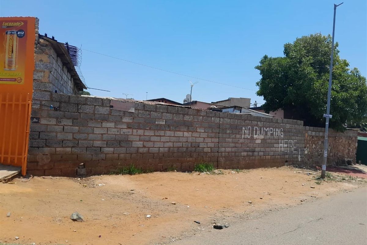 Street-level view of a vacant residential plot featuring a perimeter masonry boundary wall with razor wire and an unpaved verge.