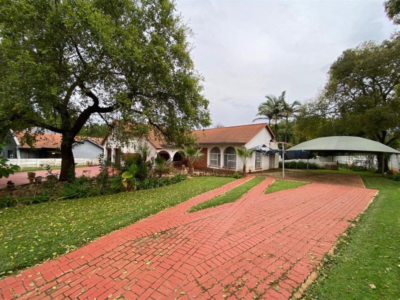 Front view of a single-story residence featuring a paved driveway, arched windows, tiled roof, and a covered carport set within a mature garden.