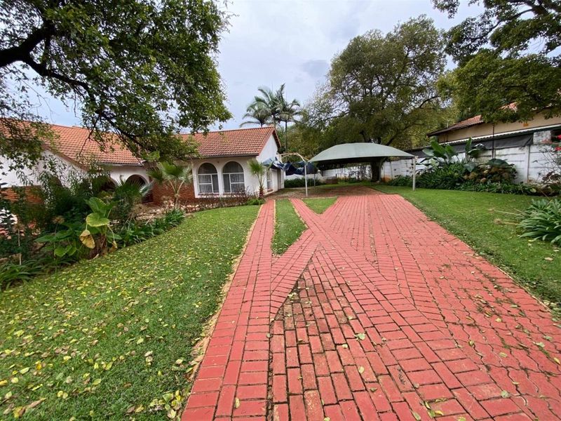 Front exterior view of the property featuring a brick driveway, carport, and established garden with mature trees.