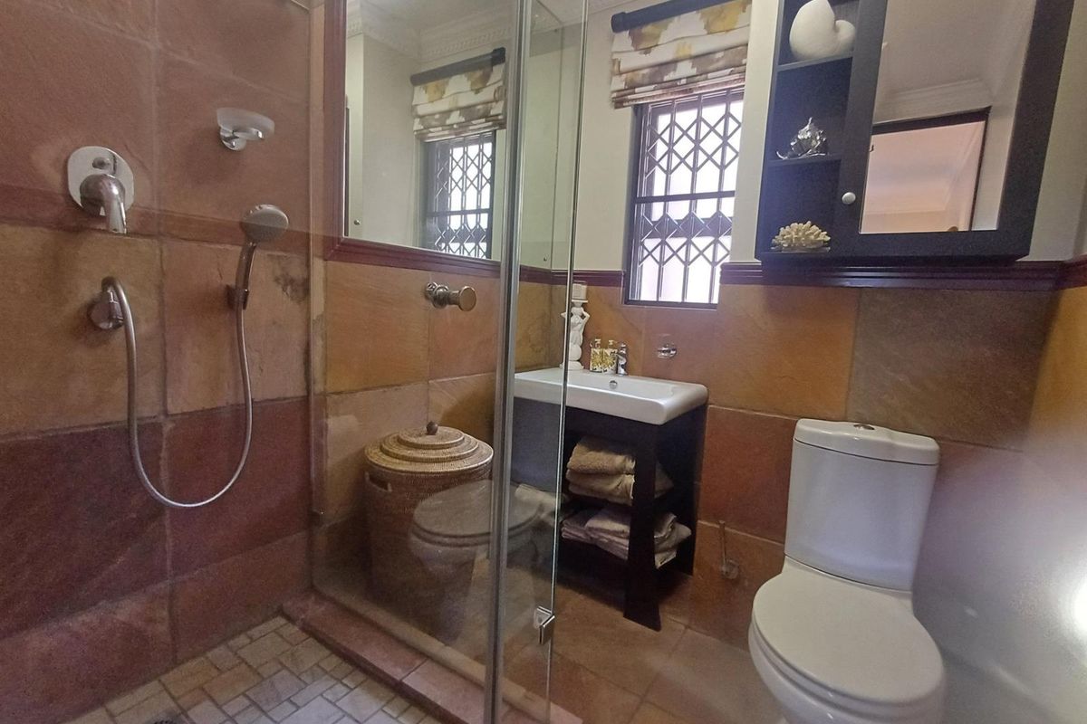 A compact bathroom featuring stone wall tiling, a glass-enclosed shower, a vanity basin, and a toilet, with security bars on the window.
