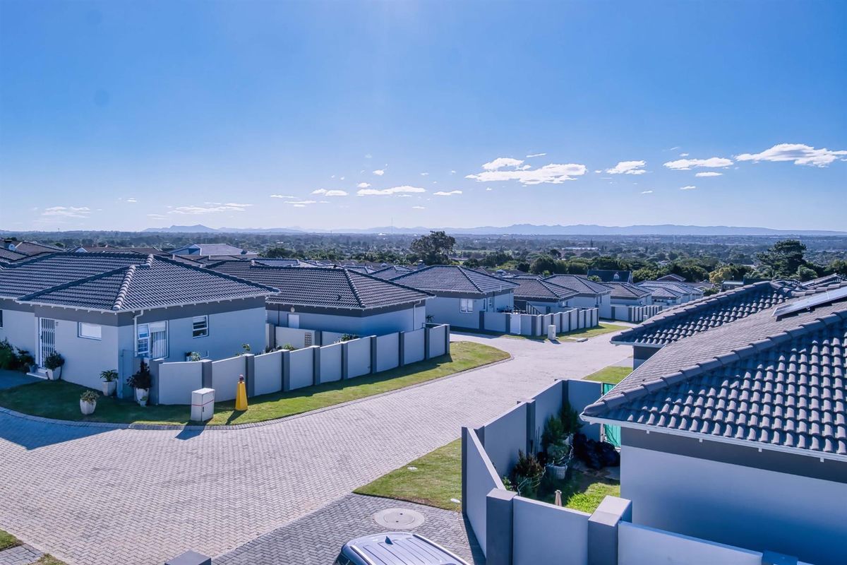 An elevated aerial view of a modern townhouse estate featuring uniform tiled roofs, paved driveways, and secure boundary walls under a clear blue sky.