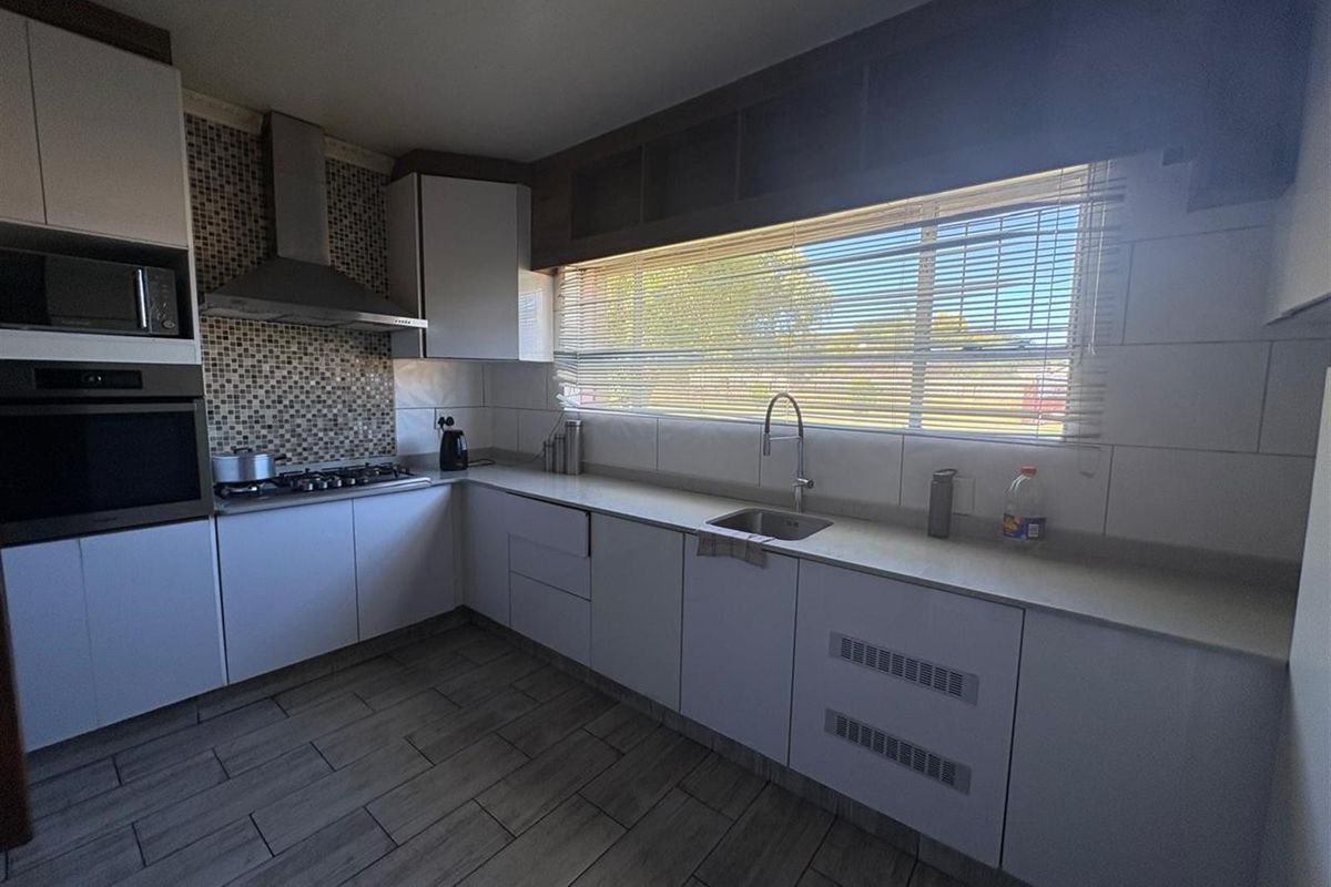 Modern kitchen featuring white cabinetry, stone countertops, mosaic tile backsplash, built-in oven, gas hob, extractor fan, and large window with blinds.