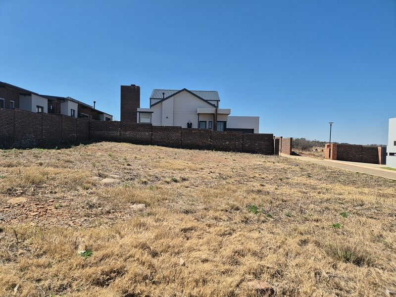 A vacant residential plot featuring a brick boundary wall and access to a paved street, situated in a developed estate setting.