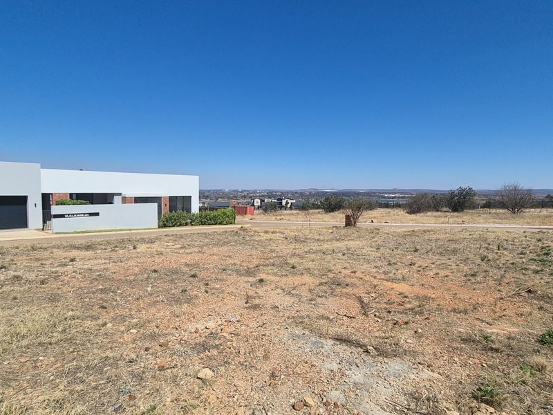 A view of a vacant residential plot with dry vegetation, adjacent to a modern building and paved driveway in a suburban estate.