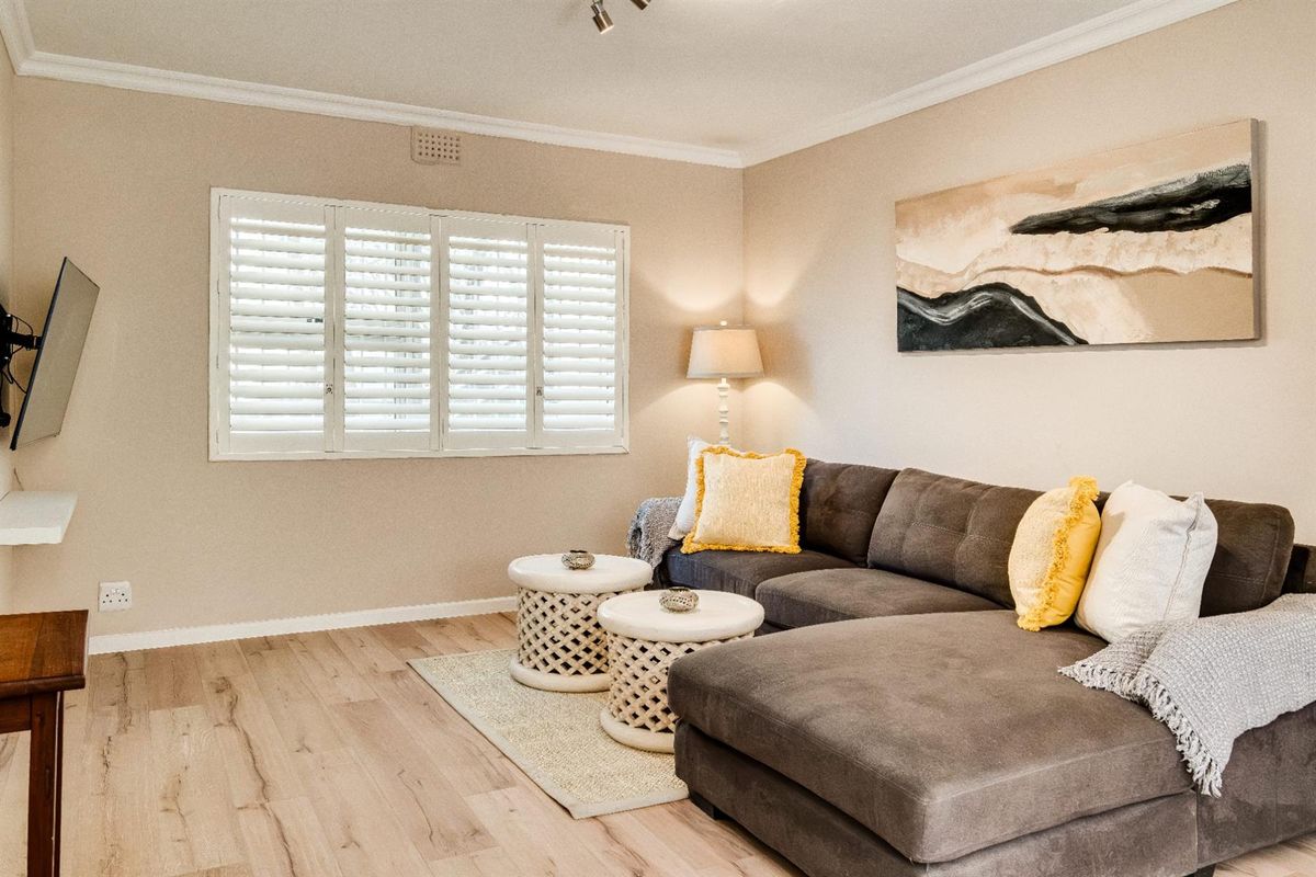 A bright and well-maintained living room featuring light wood-look flooring, crown molding, and white plantation shutters.