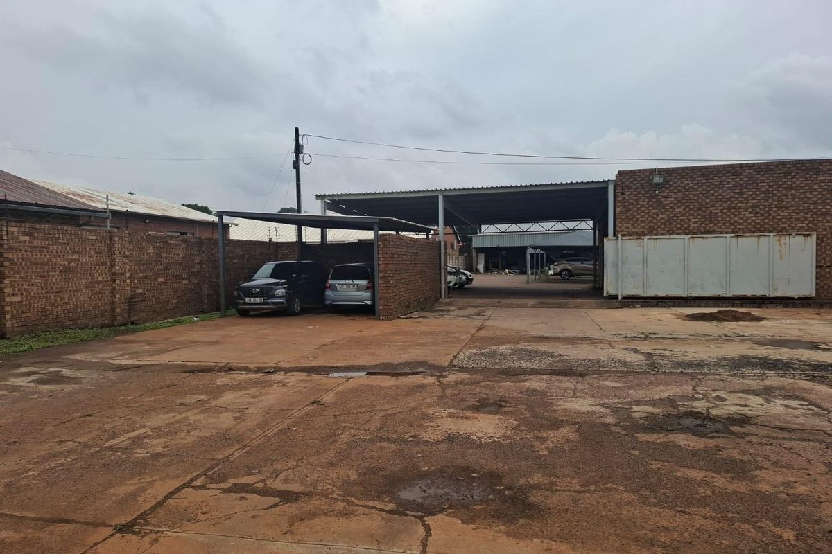 Commercial exterior yard space featuring a large concrete paved area, covered parking bays, and industrial brick structures with metal roofing.