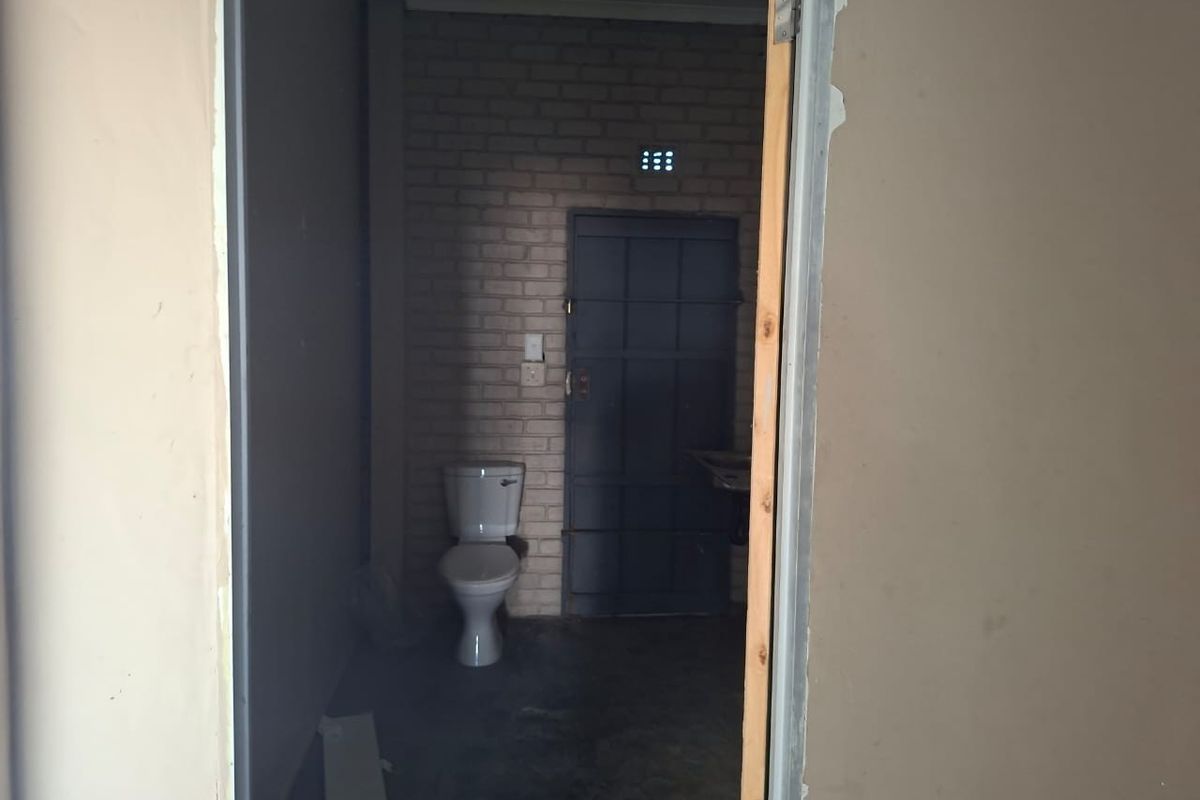 A basic, unfinished bathroom featuring an exposed brick wall, a standard toilet, and a security gate, requiring cosmetic updates and finishing touches.