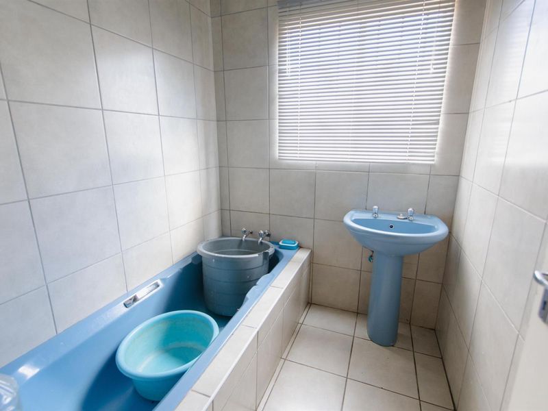A dated bathroom featuring a blue bathtub and matching pedestal basin with full-height wall tiling and a window with fitted blinds.