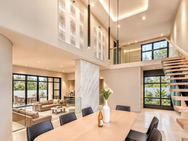 Spacious open-plan living and dining area featuring double-volume ceilings, a striking marble pillar, floating timber staircase, and expansive black-framed windows overlooking the garden.