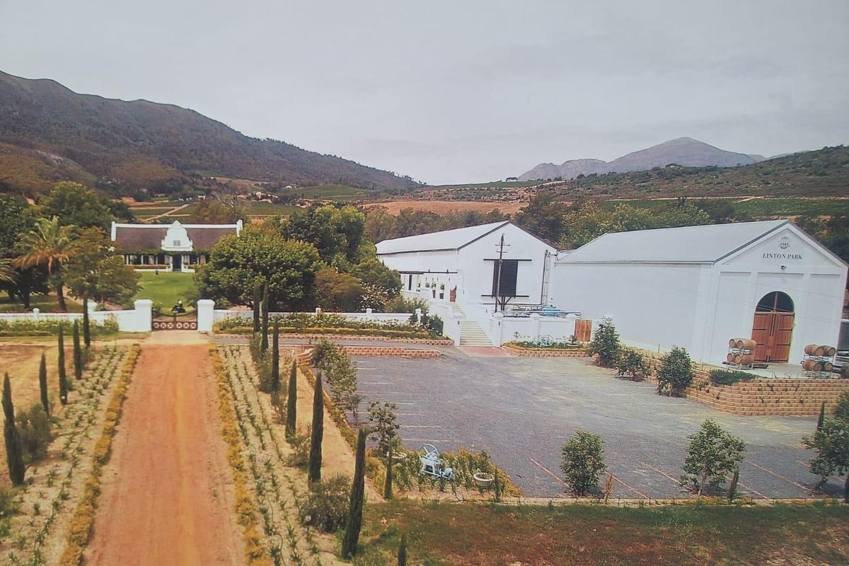 Aerial view of a farm estate featuring a gravel driveway, paved parking area, and multiple white-walled farm buildings set against a mountain landscape.