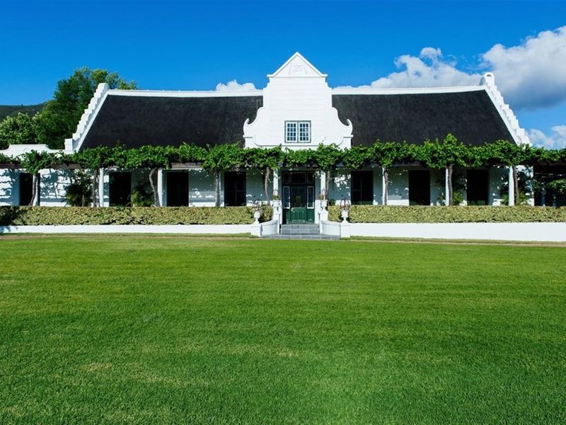 A beautifully maintained Cape Dutch style farmhouse featuring a classic thatched roof, gabled architecture, and a sprawling front lawn with a shaded pergola.