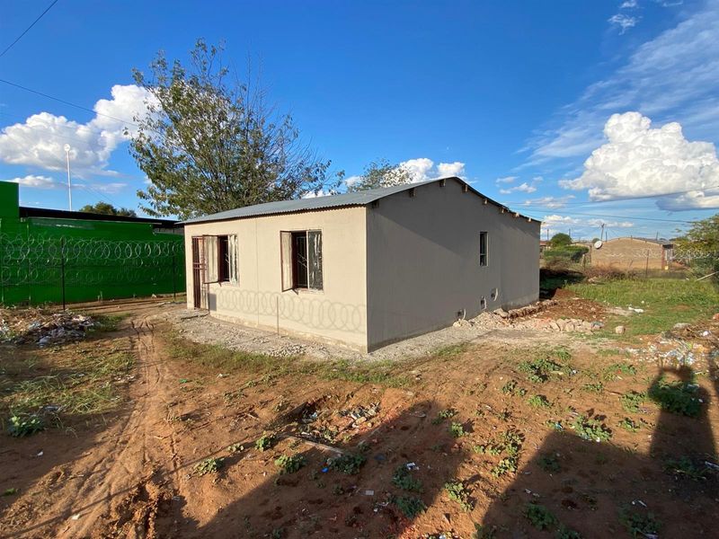 A detached single-story house featuring plastered walls and a corrugated metal roof, situated on an undeveloped plot with basic security window bars.