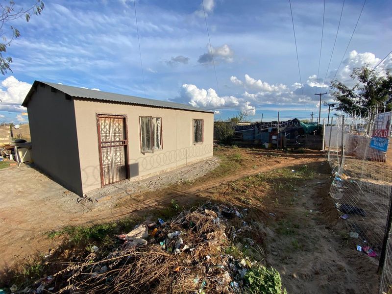 Exterior view of a modest single-story dwelling featuring a corrugated metal roof, security gate, and perimeter razor wire fencing on an undeveloped plot.