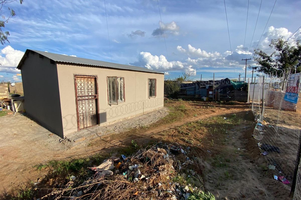 Exterior view of a modest single-story dwelling featuring a corrugated metal roof, security gate, and perimeter razor wire fencing on an undeveloped plot.