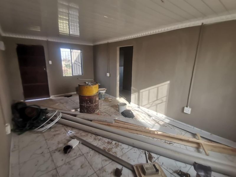 An empty living room space featuring tiled flooring, glossy ceiling panels, and security bars on the window, currently undergoing renovation or maintenance.