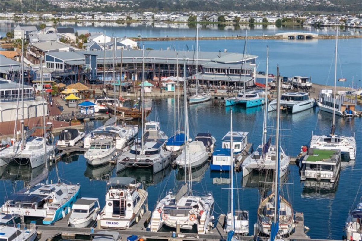 An elevated aerial view of a bustling marina featuring numerous docked yachts and catamarans, with waterfront commercial buildings and a scenic lagoon in the background.