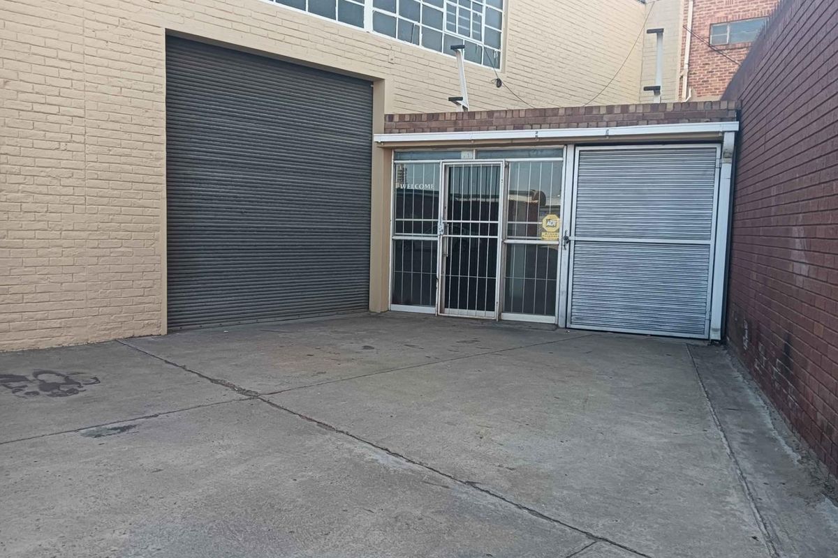 Commercial exterior view featuring a concrete courtyard, large roller shutter door, and a secure glass entrance with security gate.
