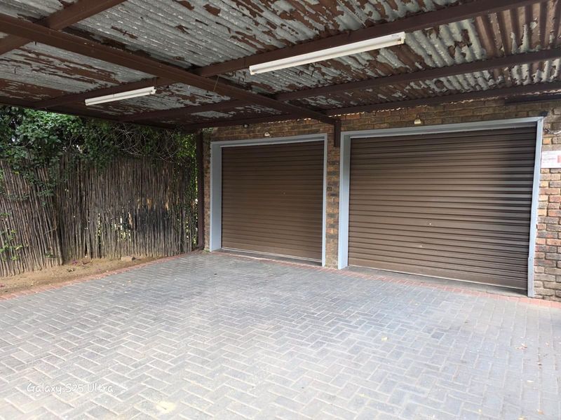 Double garage with brown roller shutter doors and a paved driveway, featuring a covered carport area with a weathered metal roof.