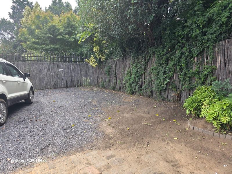 A gravel driveway area enclosed by a rustic reed fence with security wire, featuring mature trees and established perimeter greenery.