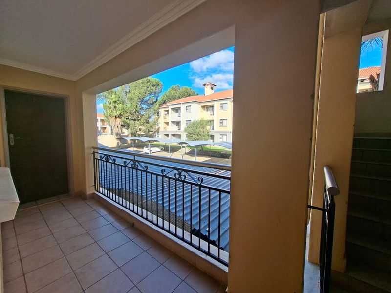 A covered balcony featuring tiled flooring and a decorative metal railing, overlooking the apartment complex grounds and parking area.