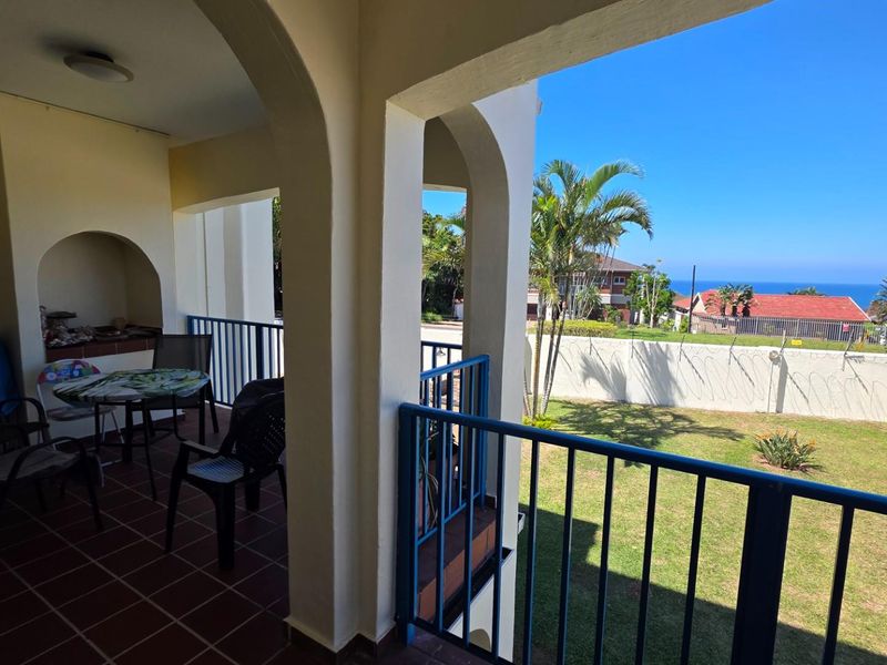 Covered balcony featuring a built-in brick braai area, tiled flooring, and metal railings, overlooking a garden with distant ocean views.