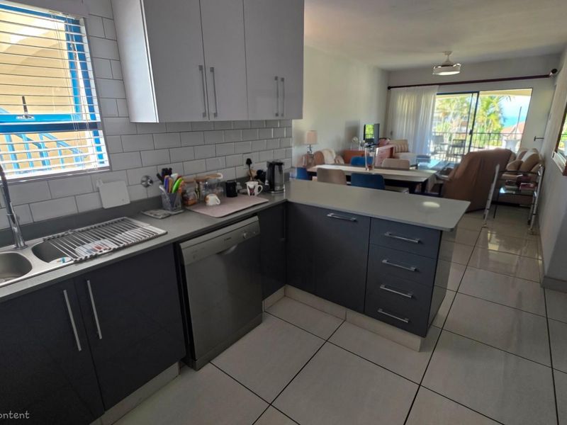 Open-plan kitchen featuring modern grey cabinetry, stone countertops, subway tile backsplash, and integrated dishwasher, flowing into the living area with balcony access.