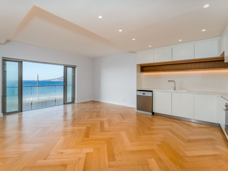 Modern open-plan kitchen and living area featuring premium herringbone timber flooring, sleek built-in cabinetry, and expansive sliding doors opening to a scenic ocean view.