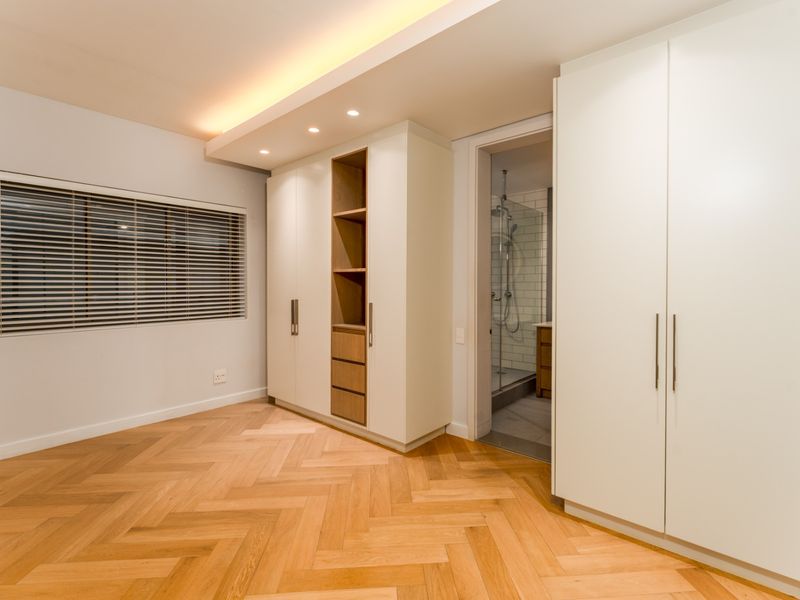 A modern bedroom featuring herringbone timber flooring, extensive built-in cabinetry, recessed ceiling lighting, and direct access to an en-suite bathroom.