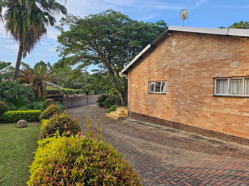 Side view of a brick house featuring a paved driveway, landscaped garden, and mature trees.