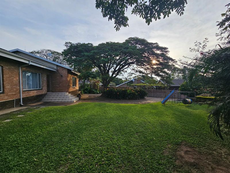Spacious rear garden featuring a well-maintained lawn, mature trees, paved patio area, and direct access to the house via tiled steps.