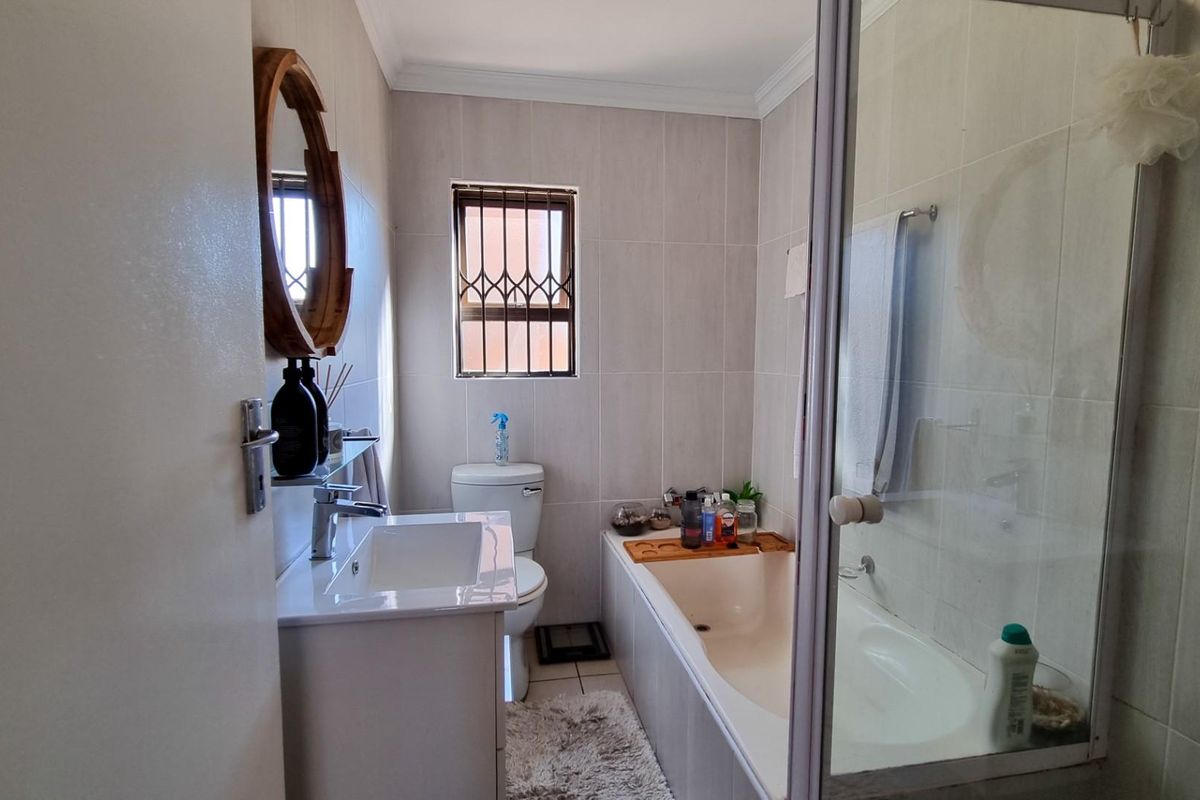 A functional bathroom featuring a built-in bathtub with shower screen, vanity basin, and toilet, finished with neutral wall tiles and a security-barred window.