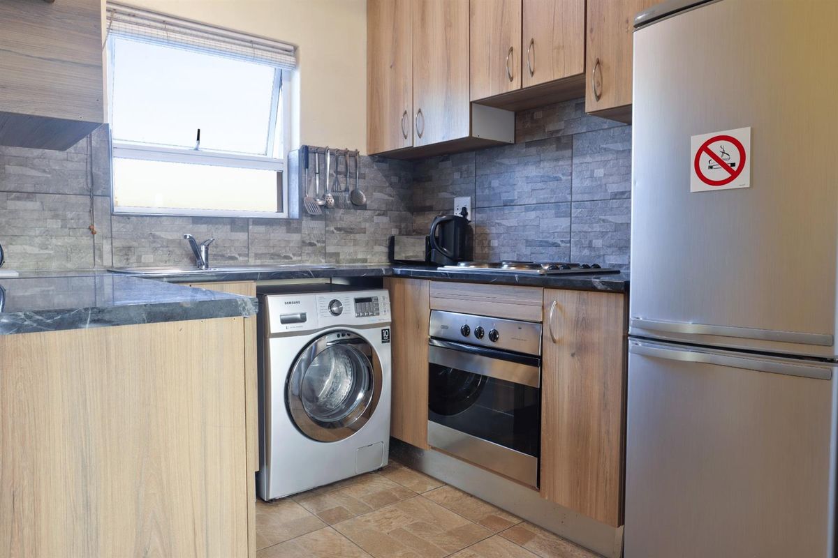 Compact kitchen featuring wood-finish cabinetry, stone-look tiled backsplash, built-in oven, hob, and integrated space for laundry appliances.