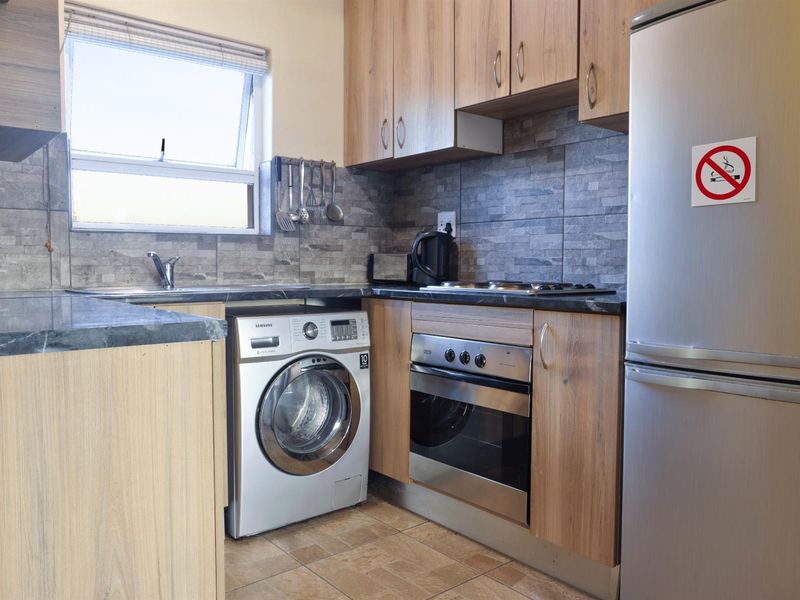 Compact kitchen featuring wood-finish cabinetry, stone-look tiled backsplash, built-in oven, hob, and integrated space for laundry appliances.
