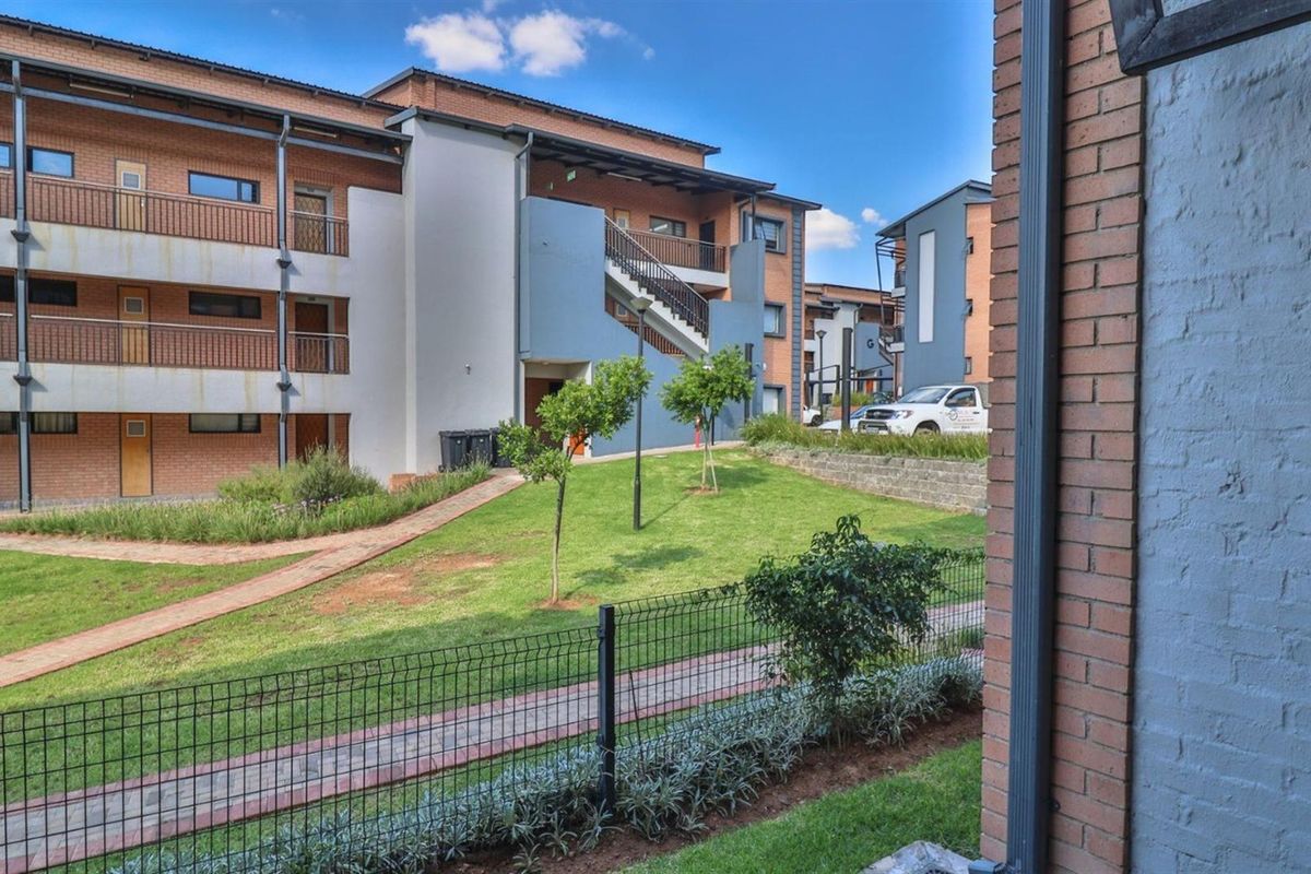 View of the apartment complex exterior featuring brick facades, balconies with metal railings, landscaped garden areas, and paved walkways.