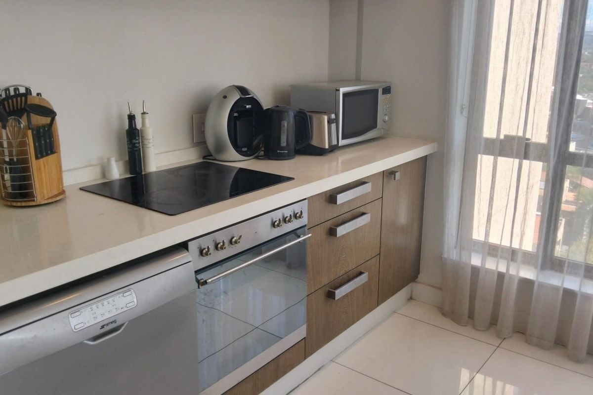 Modern kitchen featuring sleek cabinetry, stone countertops, integrated oven, electric hob, and dishwasher, with large windows providing natural light.